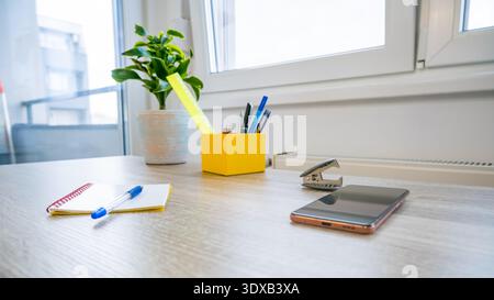 Ein moderner Arbeitsbereich mit Holzschreibtisch, grüner Topfpflanze, gelbem Stifthalter, Stiften, Haftnotizen und Smartphone. Helles natürliches Licht tritt ein Stockfoto
