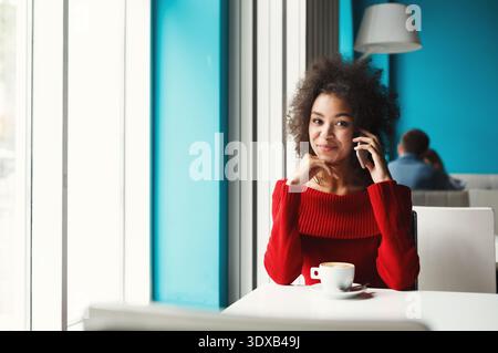 Junge Frau sitzt im Café und genießt Kaffee, während sie am Telefon in einem hellen Raum spricht Stockfoto