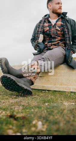 Ein Mann, der ein kariertes Hemd und eine graue Hose trägt, sitzt mit den Füßen hoch auf einer Bank. Er trägt Stiefel mit Tarnmotiven Stockfoto