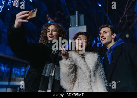 Drei Freunde machen nachts ein Selfie in stilvollen Mänteln in festlicher, urbaner Umgebung Stockfoto