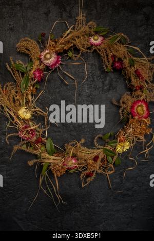 Rustikaler Kranz aus getrocknetem Stroh, grünen Blättern und bunten Blumen auf einem dunklen strukturierten Hintergrund. Natur- und Vintage-Dekoration für Zuhause, Herbst, Stockfoto
