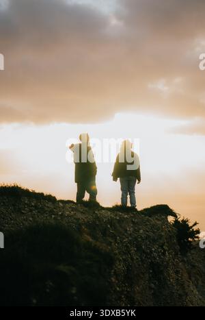 Zwei Leute stehen auf einem felsigen Hügel, einer trägt einen Hut. Die Sonne untergeht und strahlt ein warmes Leuchten über die Szene Stockfoto
