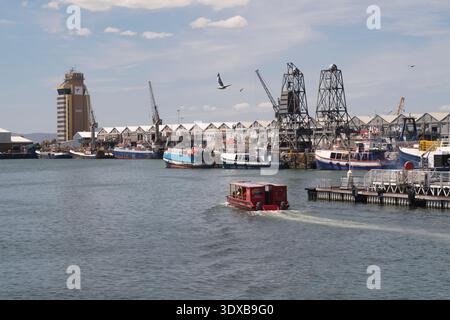 Hafen von Kapstadt mit einer roten Bootstour auf dem Kanalkanal von Kapstadt, Hop-on-Hop-off-Bootstour, die im Vordergrund und im hinteren Teil I&J vorbeifährt. Stockfoto