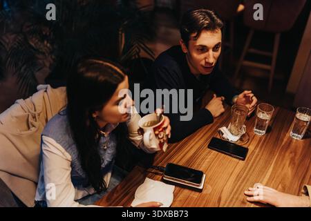 Zwei Freunde sitzen an einem hölzernen Kaffeetisch und unterhalten sich bei Getränken mit Smartphones in der Nähe Stockfoto