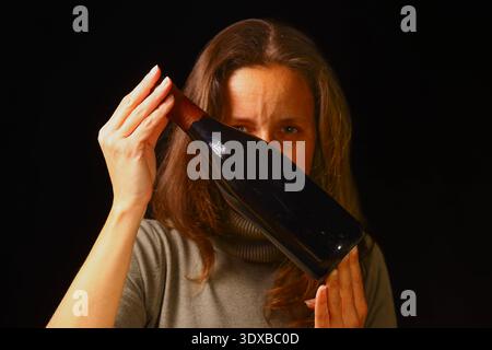 Frau mit langen Haaren hält eine Flasche Rotwein nahe an ihr Gesicht in einem dunklen Raum, die einen ruhigen Moment der Einsamkeit, emotionaler Spannung, Person hervorhebt Stockfoto