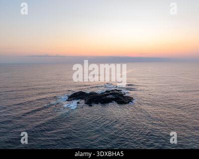 Aus der Vogelperspektive auf eine kleine, dunkle felsige Insel inmitten des Ozeans, mit weißen Wellen, die gegen ihre Kanten krachen, Sawtell, New South Wales, Australien Stockfoto