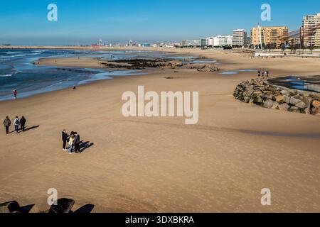 Weite Aussicht auf den Strand von Matosinhos in Portugal mit Menschen, die im Sand spazieren, die Skyline der Stadt und den Hafen von Leixões unter einem klaren blauen Himmel. Stockfoto
