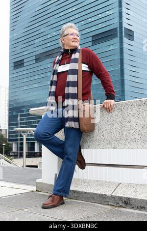Ein älterer asiatischer Mann, der auf die Betonbalustrade im städtischen plaza mit einem burgunderroten Pullover steht Stockfoto