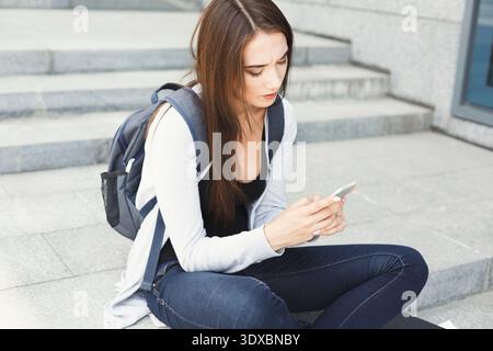 Junge Frau sitzt auf Stufen und benutzt das Telefon, während sie einen Rucksack in einer urbanen Umgebung trägt Stockfoto