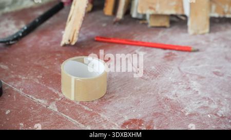 Rolle transparentes Klebeband auf staubrotem Boden mit Bleistift und Holzbrettern im Hintergrund während der Renovierungsarbeiten. Stockfoto