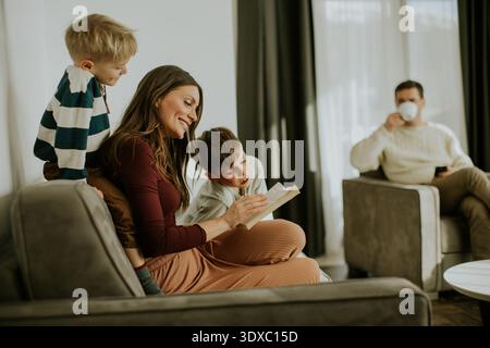 Eine Mutter liest ihren beiden Söhnen ein Buch, während sie auf einer bequemen Couch sitzt. Der Vater sitzt in der Nähe, hält einen Becher und beobachtet den Moment der Familie. Sunl Stockfoto