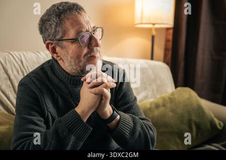 Ein reifer Mann sitzt zu Hause auf einem Sofa in einem ruhigen und reflektierenden Moment, die Hände sind gefesselt, während er ein Smartphone benutzt. Konzept von langsamem Leben, Achtsamkeit, Graben Stockfoto