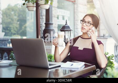 Frau, die tagsüber am Laptop arbeitet und am Telefon im Café telefoniert Stockfoto