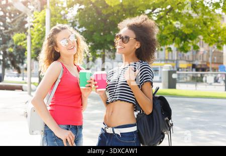 Zwei Freunde genießen an einem sonnigen Tag in einem Stadtpark einen Drink, während sie lächeln und gemeinsam Selfies machen Stockfoto
