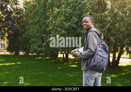 Junger Mann steht mit Rucksack im Park, hält Kaffeetasse und lächelt bei sonnigem Wetter in die Kamera Stockfoto