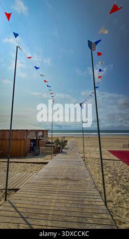 Soulac-sur-Mer, Frankreich - 4. Juni 2025. Ein hölzerner Strandgang, eingerahmt von hohen Säulen mit Girlanden aus kleinen dreieckigen Fahnen in Rot, weiß und Blau, Stockfoto