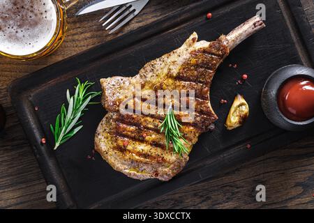 Gegrilltes Tomahawk-Steak mit Schweinefleisch auf hölzernem Schneidebrett garniert mit Rosmarinzweigen und Bier, Blick von oben. Stockfoto