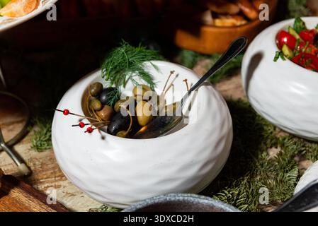 Nahaufnahme einer weißen Keramikschale gefüllt mit verschiedenen Oliven und Kapern, garniert mit frischem Dill und Cocktailpicks, mit einem Metalllöffel, der Insini aufliegt Stockfoto