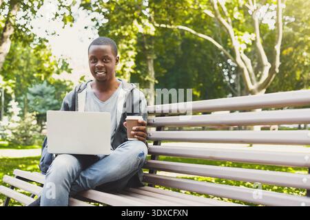 Junger Mann, der an einem Laptop im Park arbeitet, während er an einem sonnigen Tag eine Kaffeetasse hält Stockfoto