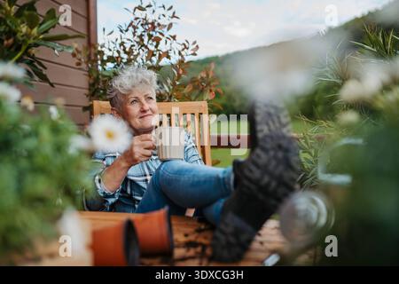 Ältere Frau mit Kaffee, die auf der Terrasse sitzt und nach der Gartenarbeit ausgeruht ist. Ältere Frau mit Kaffee, die auf der Terrasse sitzt und nach der Gartenarbeit ausgeruht ist. frühlingsgar Stockfoto