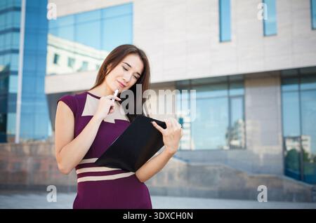 Frau im lila Kleid denkt, während sie einen schwarzen Ordner in einer modernen Stadt hält Stockfoto