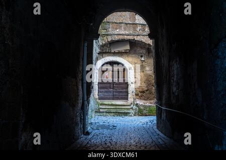 Ein malerischer Blick auf das mittelalterliche Dorf Calcata Vecchia, Latium. Das Bild umrahmt durch einen schattigen Steinbogen eine alte dunkle Holztür Stockfoto