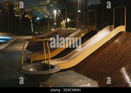 Nasser Spielplatz rutscht nachts in einen Park, beleuchtet von Straßenlaternen mit leeren Schaukeln und nass Stockfoto
