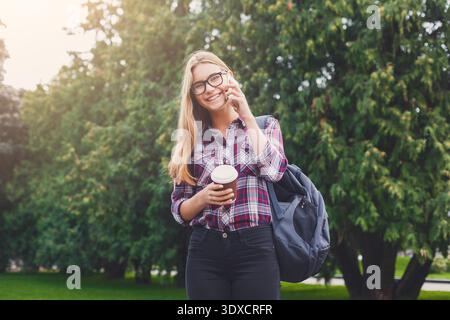 Fröhliches Mädchen spricht am Telefon, während er im Park, umgeben von Bäumen, Kaffee hält Stockfoto