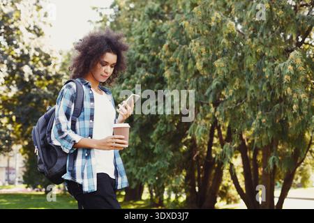 Junge Person genießt tagsüber Kaffee, während sie in einem Park mit grünen Bäumen telefoniert Stockfoto
