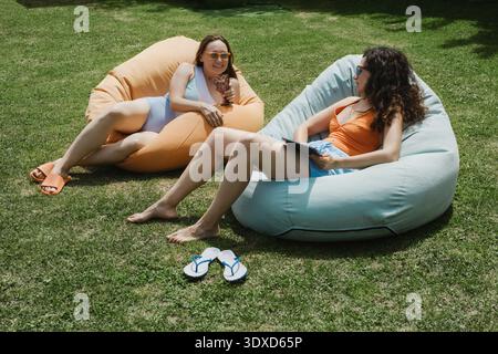 Zwei Freundinnen entspannen sich auf Sitzsack-Stühlen auf einem grünen Rasen mit einem Tablet und unterhalten sich. Sie genießen einen sonnigen Sommertag im Hinterhof während des Urlaubs. Stockfoto