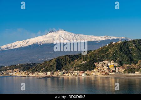 Der schneebedeckte Ätna erhebt sich über einer bezaubernden sizilianischen Küstenstadt mit farbenfrohen Gebäuden auf grünen Hügeln, die sich im ruhigen mediterranen Wasser spiegeln Stockfoto