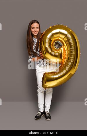 Ein Mädchen steht in einem Studio-Setting und hält einen großen goldenen Ballon in der Form der Nummer neun Stockfoto