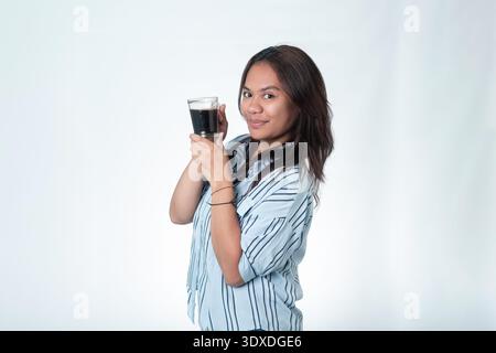 Porträt einer jungen indonesischen Frau in einem gestreiften Hemd, der Kaffee hält. Konzept für morgendliche Routine, Kaffeepause und Entspannung zu Hause oder im Büro. Stockfoto