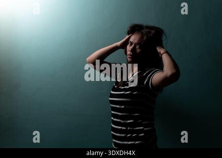 Porträt einer gestressten indonesischen Frau, die ihren Kopf hält. Filmische Aufnahmen bei schlechten Lichtverhältnissen für psychische Gesundheit, Migräne und emotionale Druckkonzepte. Stockfoto
