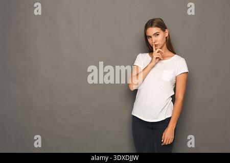 Frau in weißem Hemd macht eine Geste, um ruhig zu sein vor grauem Hintergrund mit weichem Licht Stockfoto