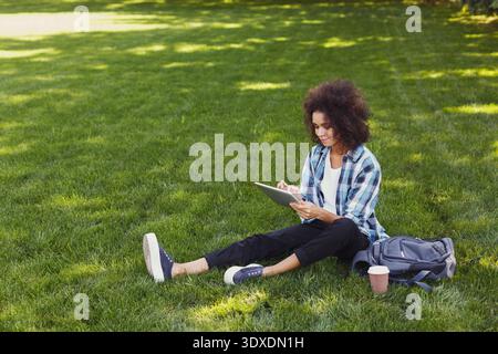 Junge Frau, die tagsüber mit einem Tablet und einer Kaffeetasse in einem Park auf Gras sitzt Stockfoto