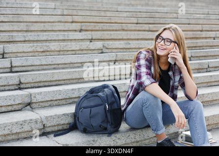 Junge Frau sitzt auf einer Treppe und spricht am Telefon in einer ungezwungenen Outdoor-Umgebung mit einem Rucksack in der Nähe Stockfoto