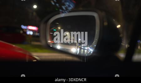 Kinoaufnahme durch einen Seitenspiegel eines Autos mit einem weißen Fahrzeug mit hellen Scheinwerfern im Verkehr, urbanem Bokeh und verschwommenem Stadtlicht im B Stockfoto