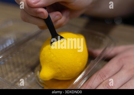Person, die eine schwarze Gabel verwendet, um ein realistisches Zitronenmousse-Dessert zu probieren, das in einem Plastikbehälter serviert wird, Nahaufnahme von lebendigem gelbem Zitrusgebäck Stockfoto
