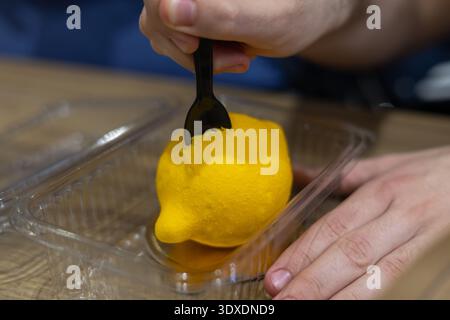 Zitronenmousse-Dessert in einem Plastikbehälter, das mit einem schwarzen Löffel probiert wird, mit realistischer Zitrusstruktur und kreativer Gebäckkunst in cl Stockfoto