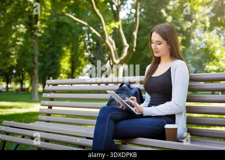 Junge Frau sitzt am sonnigen Tag auf der Bank im Park mit Tablet Stockfoto