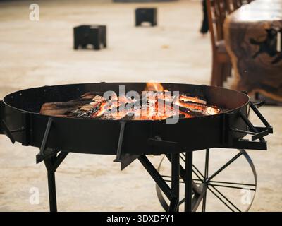 Eine schwarze Metallschale mit einem Rad ist mit brennendem Holz gefüllt. Die Schüssel steht auf einem Tisch und hat ein paar Stühle um sie herum. Die Szene ist warm und einladend Stockfoto