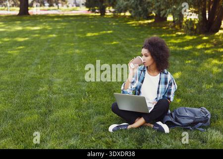 Junge Person genießt Kaffee, während sie an einem Laptop in einem grasbewachsenen Außenbereich arbeitet Stockfoto