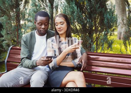 Ein Paar sitzt auf einer Bank in einem Park, während er sein Handy in sonniger Umgebung benutzt Stockfoto