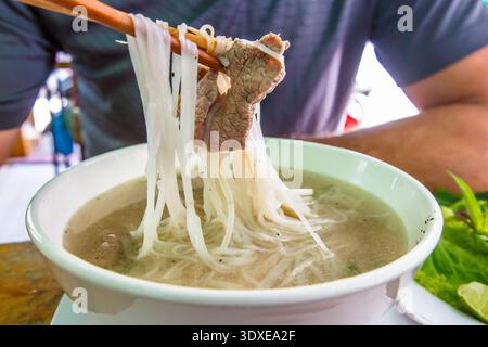 Ein Mann isst traditionelle vietnamesische Rindersuppe Pho Bo mit Essstäbchen. Street Food. Eine Schüssel Pho mit Nudeln und Rindfleisch essen. Mann hält Essstäbchen Stockfoto