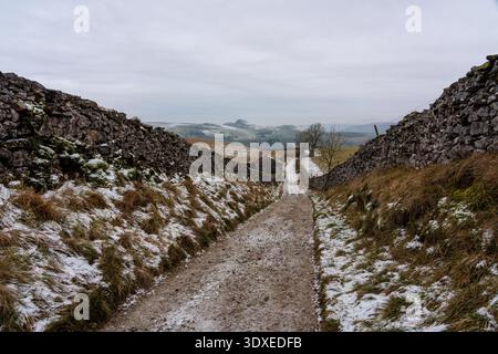 Der frostbedeckte Kiesweg führt zwischen hohen Trockenmauern zu einer entfernten Steinscheune und schneebedeckten Hügeln in den Yorkshire Dales Stockfoto