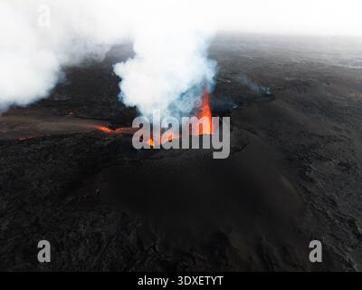 Aus dem dunklen Krater des Vulkans bricht feurige Lava aus der Vogelperspektive auf, im Kontrast zum weißen Rauch vor der einsamen Landschaft, Grindavik, Island. Stockfoto