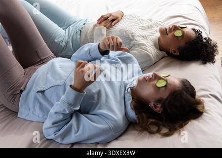 Verschiedene Freundinnen liegen mit Gurkenscheiben nebeneinander auf dem Bett, tragen Hoodie und Pullover Stockfoto