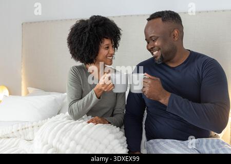 Afroamerikanisches Paar, das graue Keramikbecher hält und im Schlafanzug auf dem Bett sitzt Stockfoto