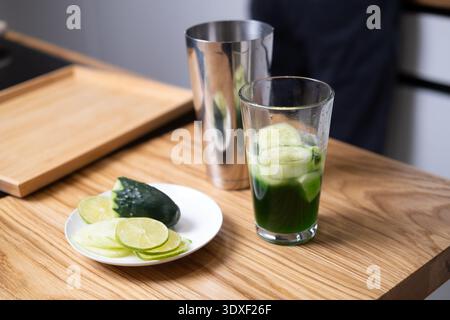 Mischen von Glas mit grüner Flüssigkeit und Gurkenscheiben auf Holztisch neben Metallschüttler und Kalk. Nachhaltige pflanzliche Getränke, umweltfreundlich Stockfoto